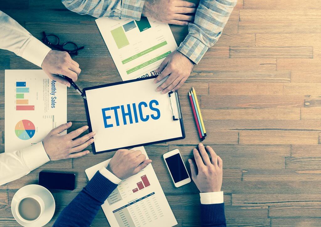 Photo of people's hands on a table looking at a clipboard with the word 'Ethics' on it