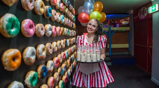 Photo of a girl holding a tray full of donuts next to a wall of donuts and balloons int he background