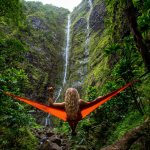 Photo of a girl with long wavy blonde hair sitting in a hammock looking at a waterfall in the forest