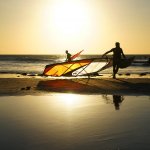 Photograph of three windsurfers coming in from the sea with their boards at sunset