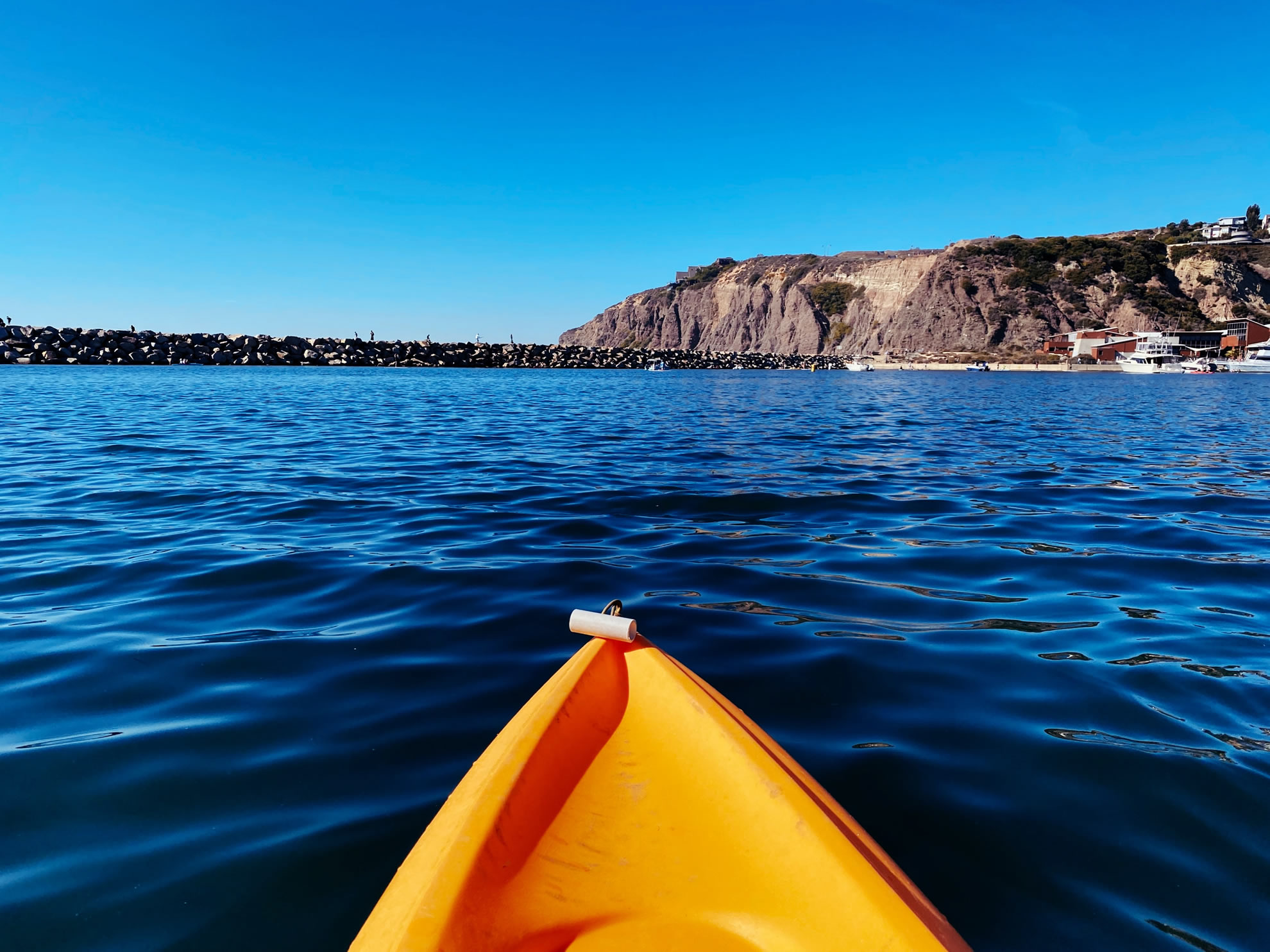 Photo of the front of an orange kayak on the ocean with a blue sky and rocks ahead