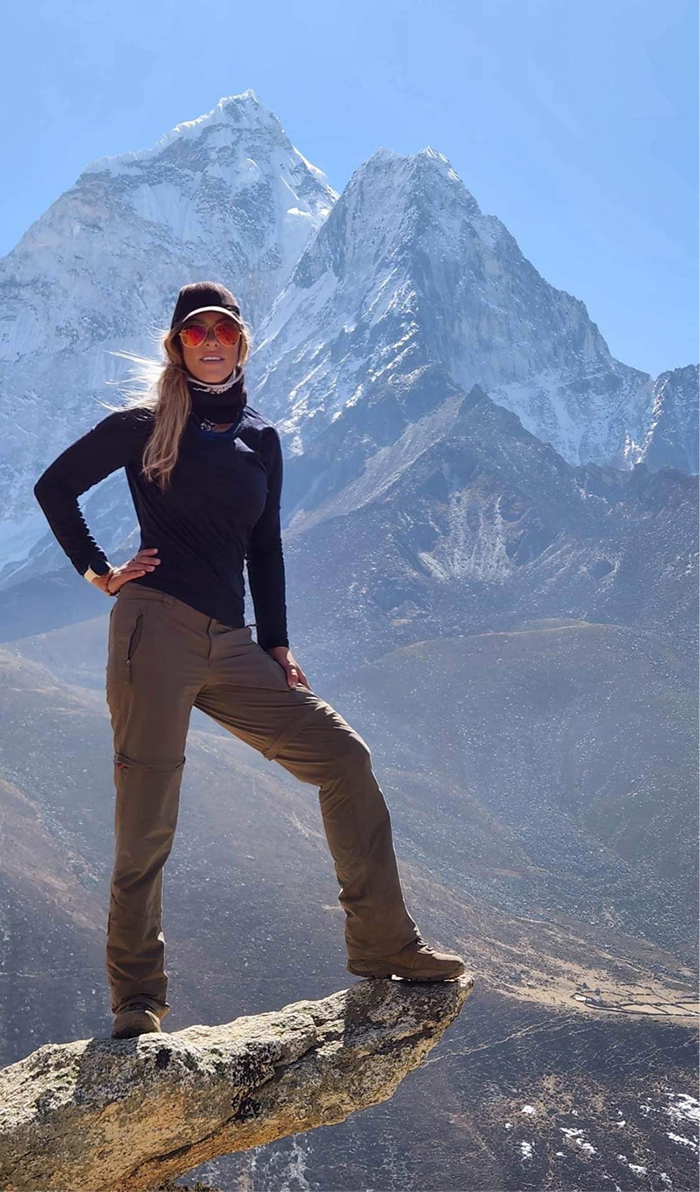 Photo of Alex Sieniarski wearing hiking gear standing on a precipice with mountains in the background.
