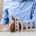 Person holding their hand out to stop wooden blocks knocking down more wooden blocks (like dominos), and the word 'RISK' is written on the one in the middle.