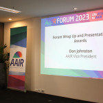 Photo of Don Johnston delivering the closing speech at the 2023 AAIR Forum. There is a pull up banner and PowerPoint slide up on stage behind him.