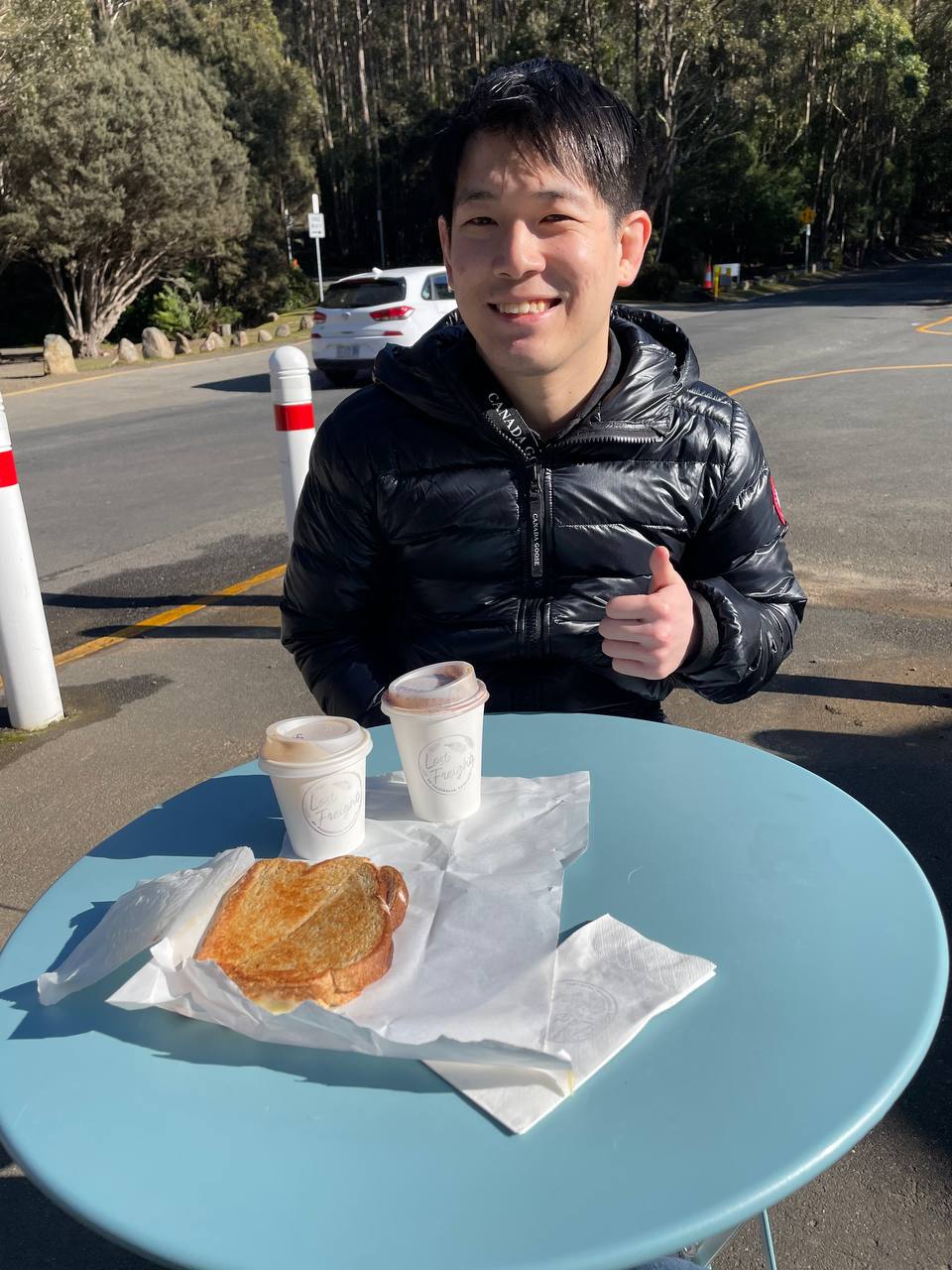 Photo of Brenwin Ang wearing a black coat sitting at a table with a toasty and two takeaway coffee cups on it. He's smiling and giving the thumbs up.
