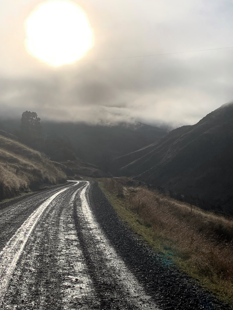 Photo of a wet gravel road, a grey wintery sky, and a sun trying to shine through the clouds. The road leads through gloomy mountains.