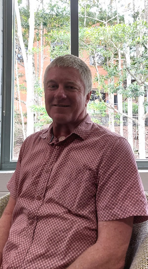 Photo of Don Johnston wearing a red shirt sitting inside with trees out the window behind him.