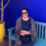 Photo of Francesca Casal Sitting on a teal coloured bench in front of a deep cobalt blue wall next to a pot plant in a bright yellow planter.