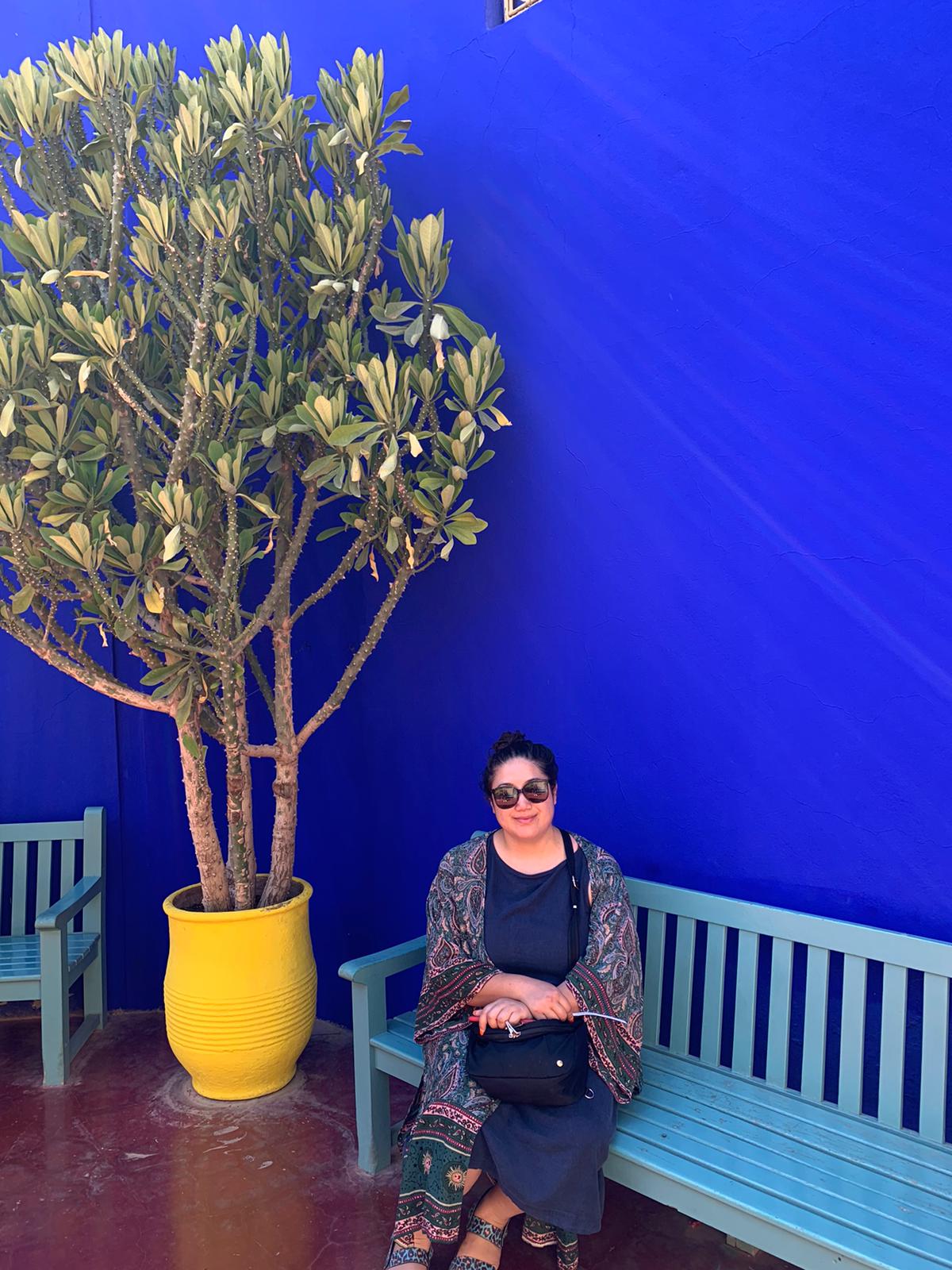 Photo of Francesca Casal sitting on a teal coloured bench in front of a deep cobalt blue wall next to a pot plant in a bright yellow planter.